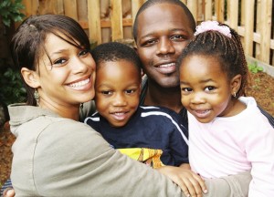 A photograph of a black family. The mother and father are hugging their young boy and girl in front of a wooden fence. Everyone is smiling.