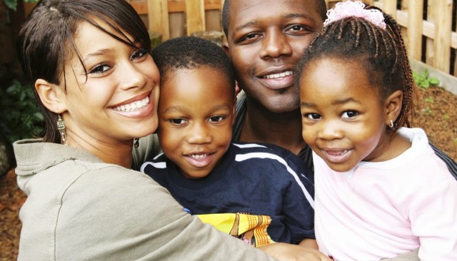 A photograph of a black family. The mother and father are hugging their young boy and girl in front of a wooden fence. Everyone is smiling.