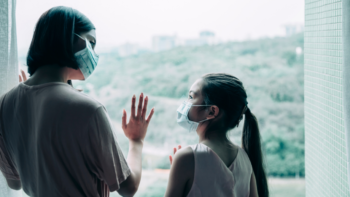 Woman wearing mask with hand on window looks down at female child, also wearing mask