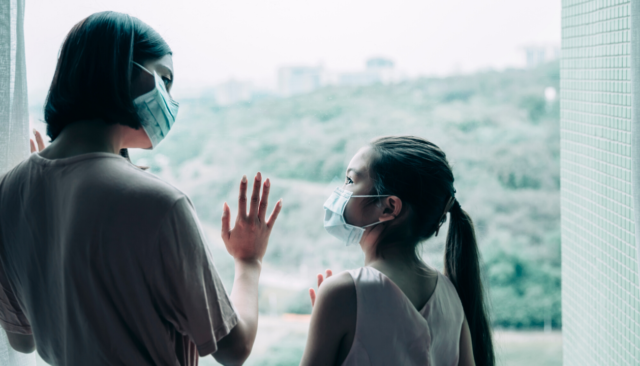 Woman wearing mask with hand on window looks down at female child, also wearing mask